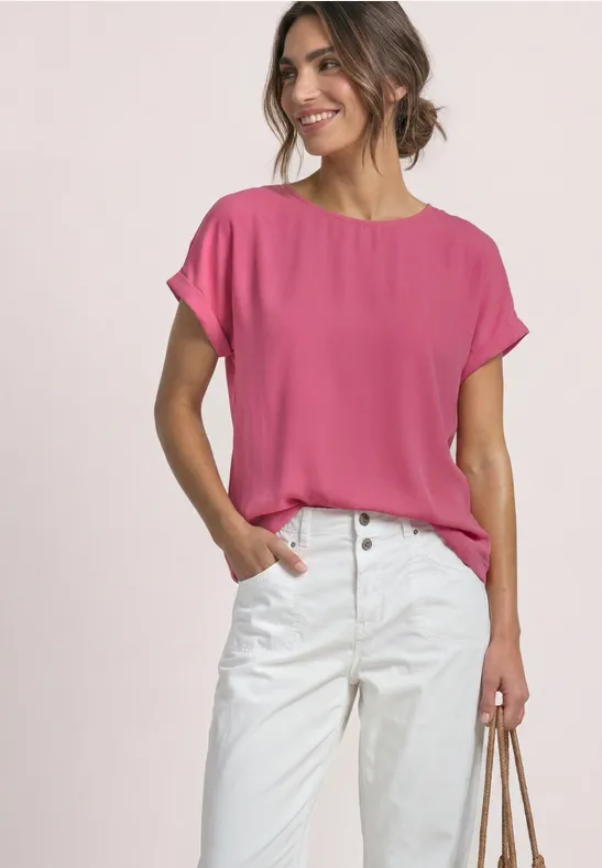 Frau trägt pinkes kurzärmeliges Oberteil und weiße Hose, lächelt, Hand in Tasche, lässiger Look.