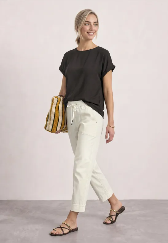 Frau in schwarzem Shirt, hellem Bundfaltenhose und Sandalen mit gestreifter Tasche, lächelnd spazierend.
