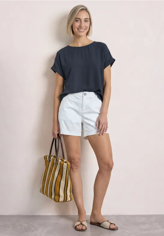 Eine lächelnde Frau in dunkelblauem Shirt, weißen Shorts und Sandalen trägt eine gestreifte Tasche.
