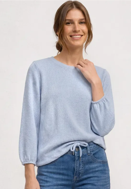Person trägt hellblauen Strickpullover mit 3/4-Ärmeln und Bindeband, kombiniert mit blauen Jeans.