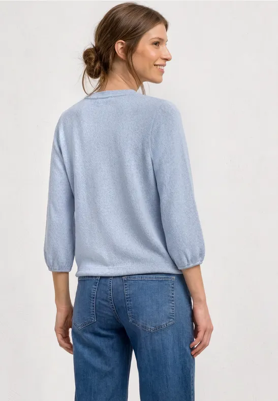 Von hinten aufgenommene Frau mit hellblauem Pullover und blauen Jeans vor neutralem Hintergrund.