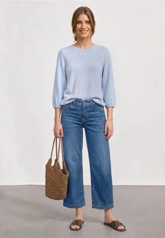 Eine Frau trägt hellblauen Pullover, weite Cropped-Jeans, Basttasche und flache Sandalen vor neutralem Hintergrund.