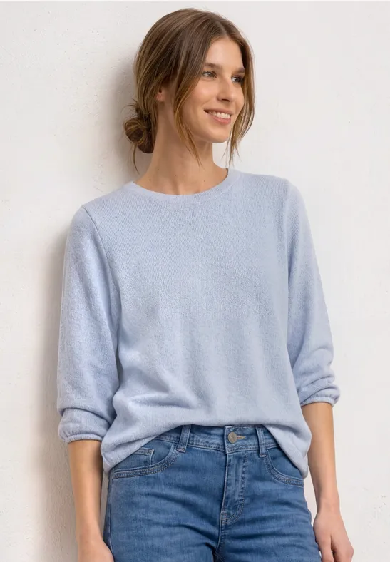 Dame in hellblauem Pullover und blauen Jeans vor neutralem Hintergrund.