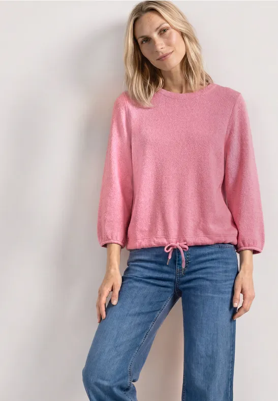 Eine blonde Frau trägt einen rosa Pullover und blaue Jeans vor hellem Hintergrund.