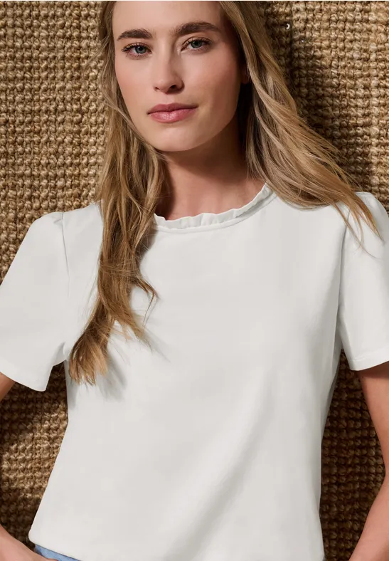 Jonge vrouw met lange blonde haren draagt een wit T-shirt met korte mouwen en ruches aan de hals voor een gevlochten achtergrond.