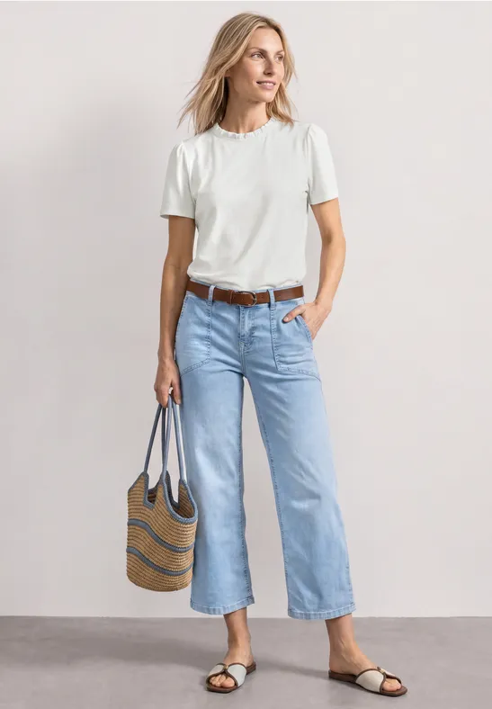 Vrouw in wit T-shirt en lichtblauwe jeans met gestreepte tas en platte sandalen tegen neutrale achtergrond.