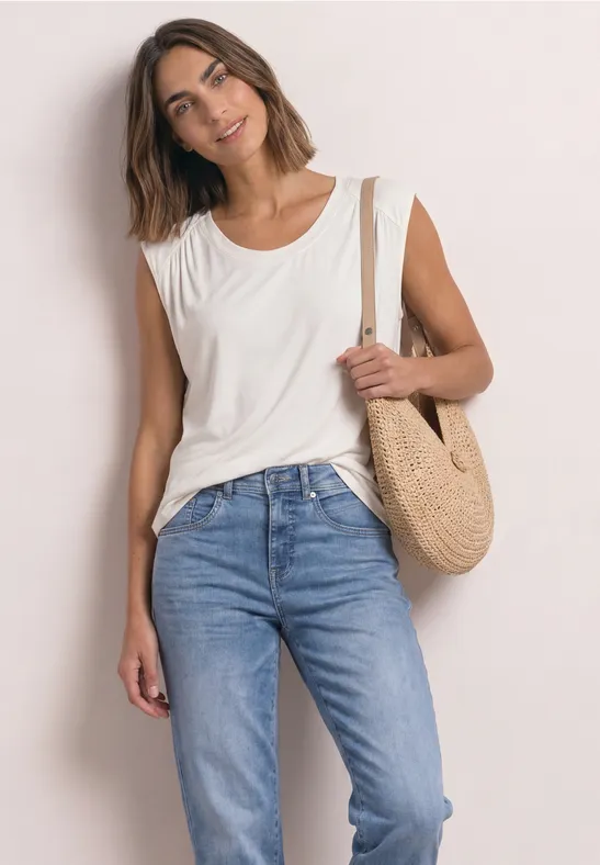 Junge Frau in weißem Top und blauer Jeans mit runder Strohtasche an heller Wand