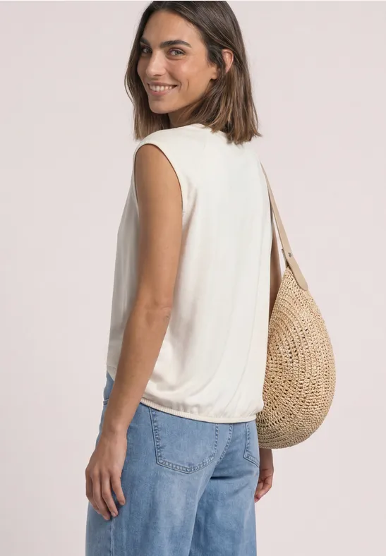 Frau in weißem ärmellosem Top und Jeans, mit Strohtasche, seitlich lachend abgelichtet.