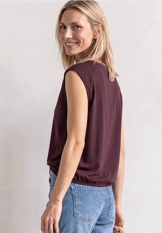 Blondynka w bezrękawniku i dżinsach, uśmiecha się przez ramię.