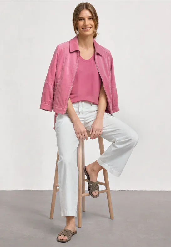 Persoon zit op kruk, draagt roze jas over roze top, witte wijde broek en sandalen.
