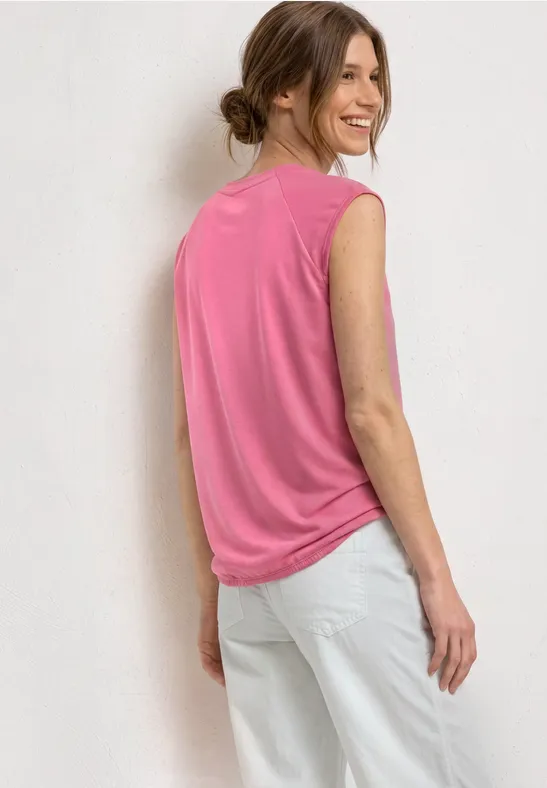 Een vrouw staat zijwaarts tegen een witte muur, draagt een roze mouwloos shirt en een witte broek.