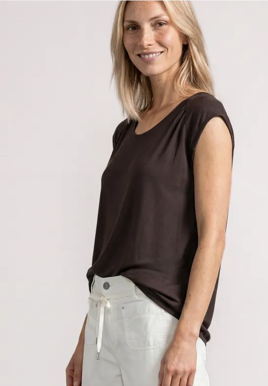 Glimlachende vrouw met bruin topje en witte broek voor een lichte achtergrond.