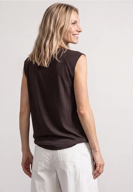 Vrouw van achteren, glimlachend over haar schouder kijkend, zwarte mouwloze top en witte broek.
