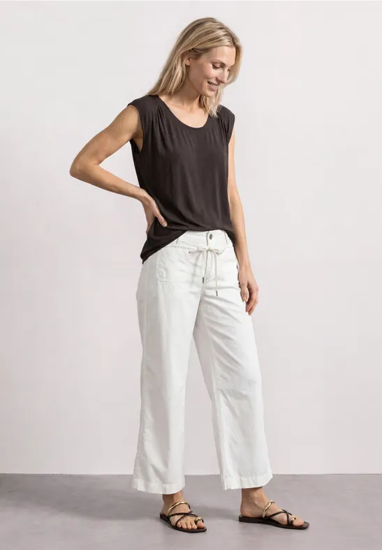 Vrouw glimlacht in zwart mouwloos top, witte wijde broek en zwarte sandalen, zijaanzicht.