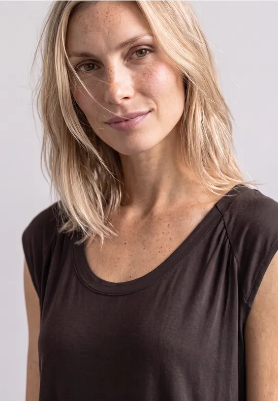 Portret van een blonde vrouw met sproeten, licht glimlachend in een donker topje voor een lichte achtergrond.