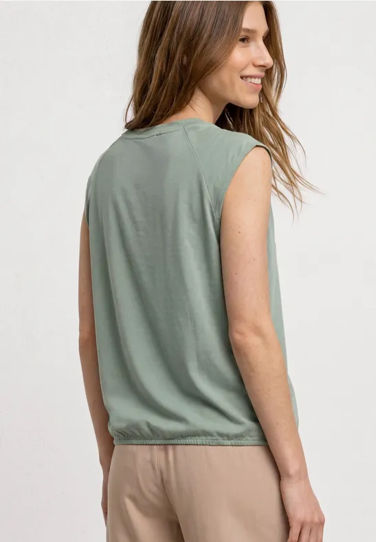 Eine Frau trägt ein ärmelloses grünes Top und bequeme beige Hosen, steht seitlich und lächelt.