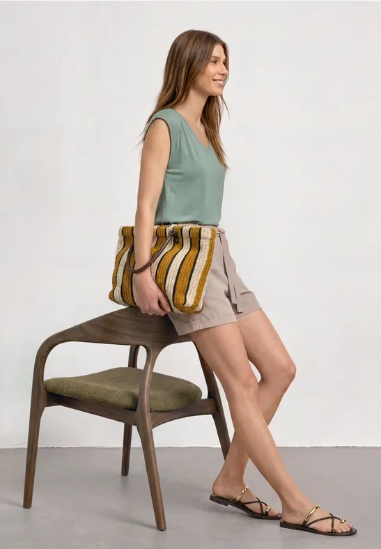 Eine Frau lehnt an einem Stuhl, trägt ein grünes Top, beige Shorts und Sandalen, hält eine gestreifte Tasche.