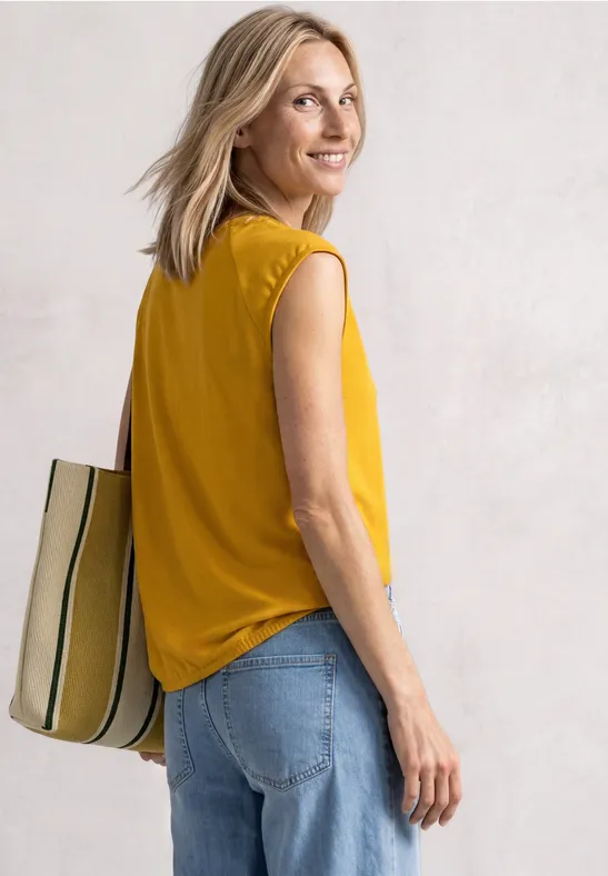 Une femme en haut jaune et jean, portant un sac rayé, sourit par-dessus son épaule.