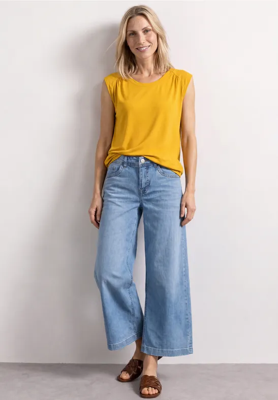 Femme en top sans manches jaune et jean large, sandales marron, devant un mur épuré.