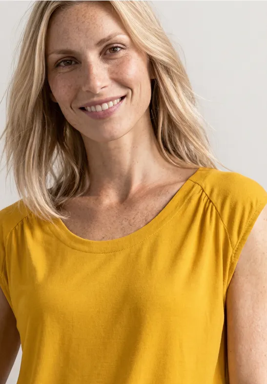 Femme blonde souriante en haut jaune devant un fond clair.