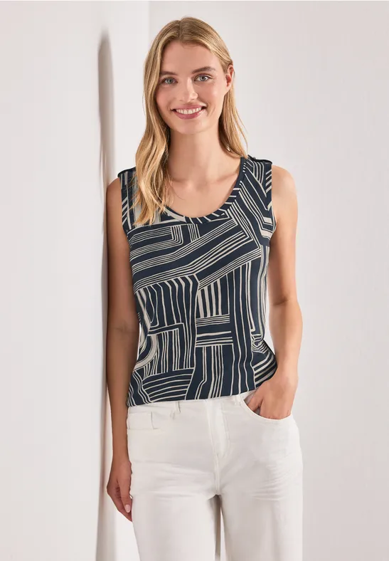 Eine lächelnde Frau in einem ärmellosen, geometrisch gemusterten Top und weißen Hosen lehnt an der Wand.