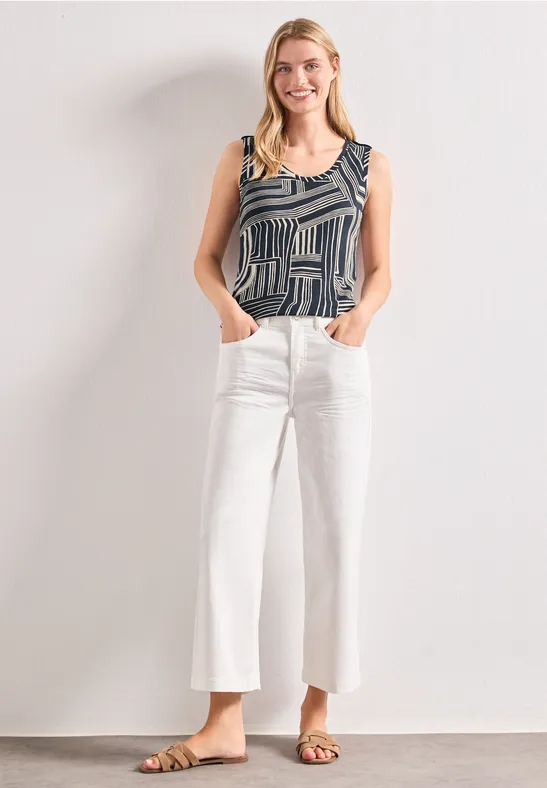 Frau in gemustertem Top, weißen weiten Cropped-Jeans und Sandalen steht mit Händen in den Taschen vor weißer Wand.