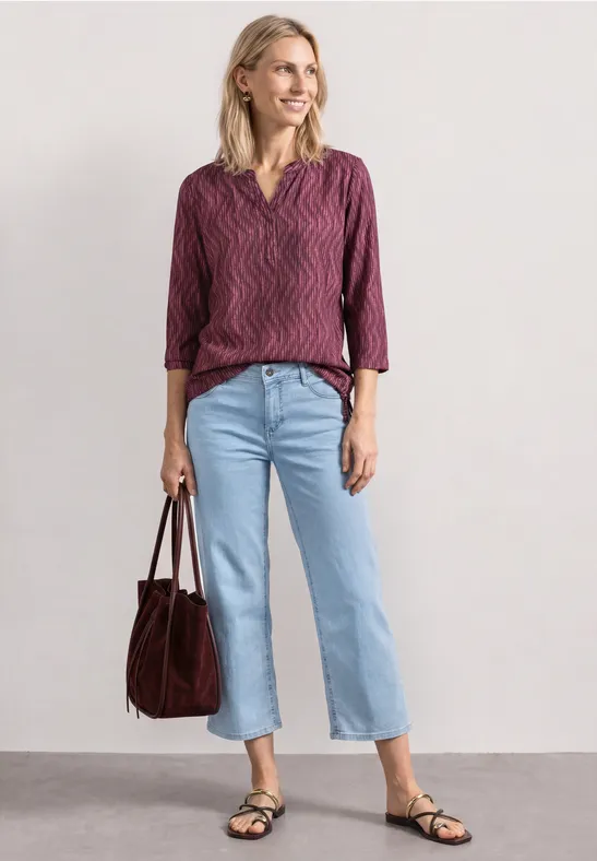 Frau mit purpurroter Bluse, hellblauen Jeans, schwarzen Sandalen und brauner Tasche vor neutralem Hintergrund