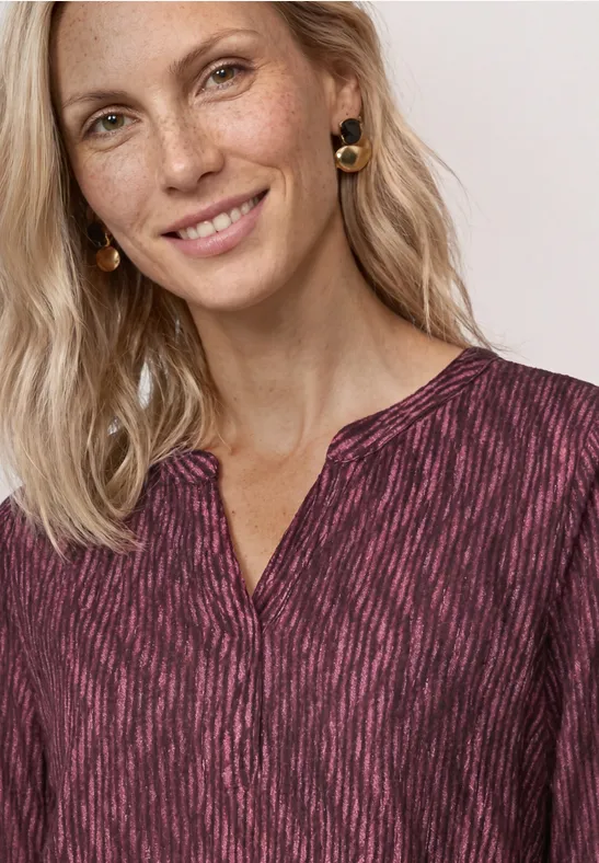 Lächelnde blonde Frau mit zarten Sommersprossen, trägt gemustertes rosafarbenes Oberteil und goldene Ohrringe.