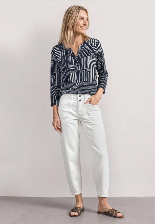 Frau in gemustertem Langarmshirt, weißen Jeans und braunen Sandalen vor neutralem Hintergrund.