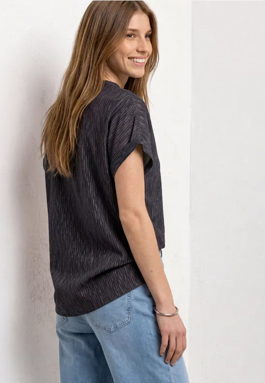 Eine lächelnde Frau in schwarzem T-Shirt und Jeans lehnt an einer Wand und blickt über die Schulter.