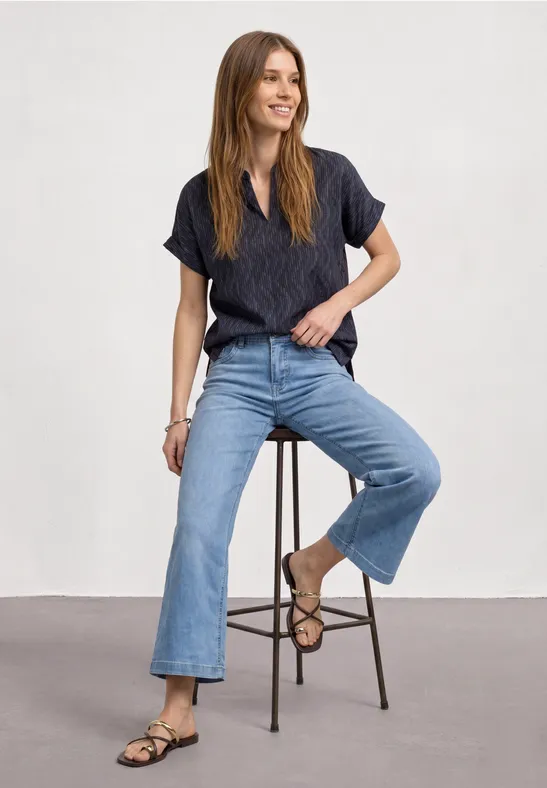 Frau sitzt lächelnd auf Hocker, trägt dunkelblaues Shirt, weite Jeans und Sandalen vor hellgrauem Hintergrund.