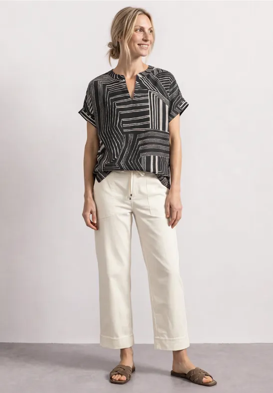 Frau trägt gemustertes schwarzes Shirt, helle Cropped-Hose und Sandalen, lächelnd stehend.