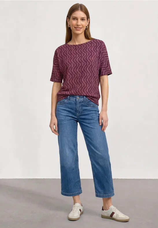 Frau in kurzärmeligem, gemustertem Shirt, weitem Jeans-Crop und weißen Sneakern vor neutralem Hintergrund.