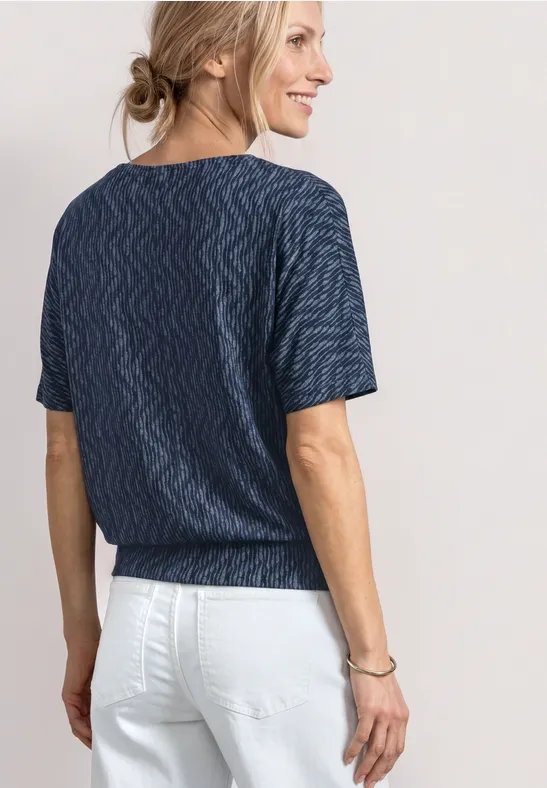 Frau von hinten mit dunkelblauem, wellenmusterigem T-Shirt und weißen Hosen, Haare zu einem lockeren Dutt gebunden.