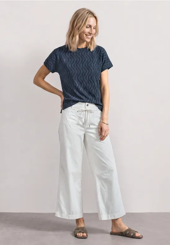 Vrouw staat voor neutrale achtergrond, draagt blauw gestreept T-shirt, witte wijde broek en sandalen.