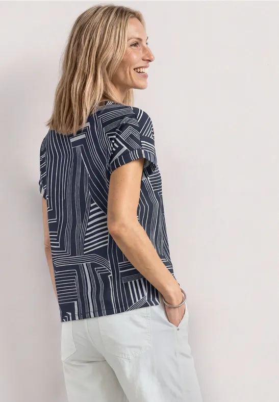 Vrouw in blauw patroon-t-shirt en witte broek, zijaanzicht tegen lichte achtergrond.