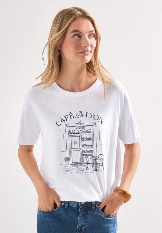 Femme en t-shirt blanc illustré « Café de Lyon », jean, cheveux lâchés, posant devant un mur clair.