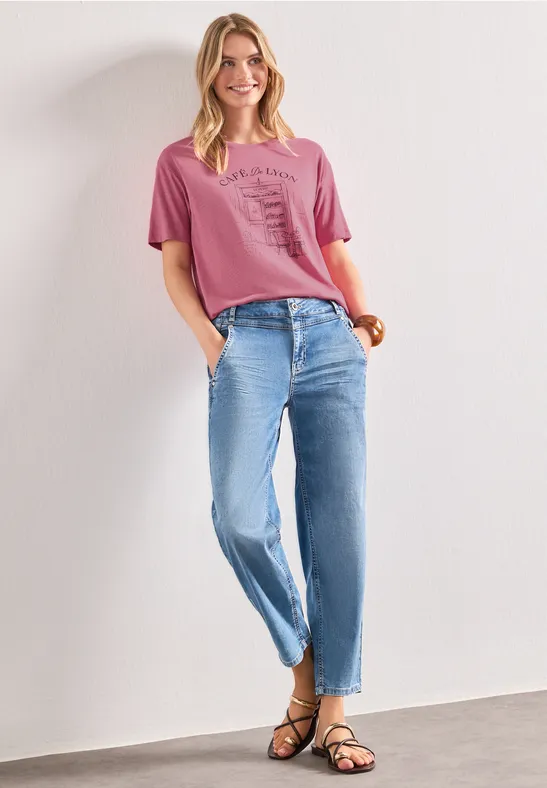 Femme en t-shirt rose, jean bleu décontracté et sandales noires, debout les mains dans les poches.