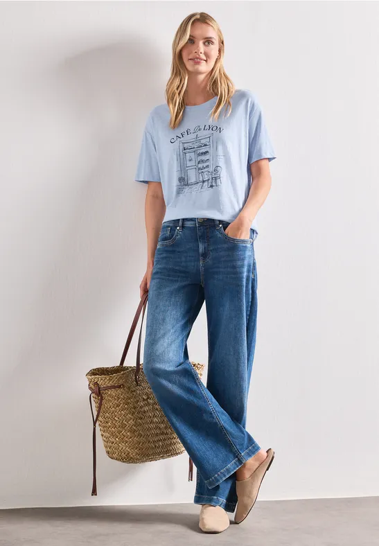 Femme au visage flouté porte un t-shirt bleu clair, jean large, sac en paille et mules.