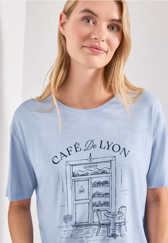 Femme porte un t-shirt bleu clair illustré 'Café de Lyon' avec table et chaise.