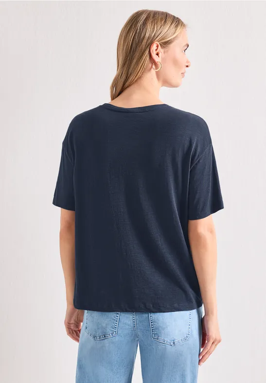 Frau steht mit dem Rücken zur Kamera, trägt dunkelblaues T-Shirt und helle Jeans vor weißer Wand.