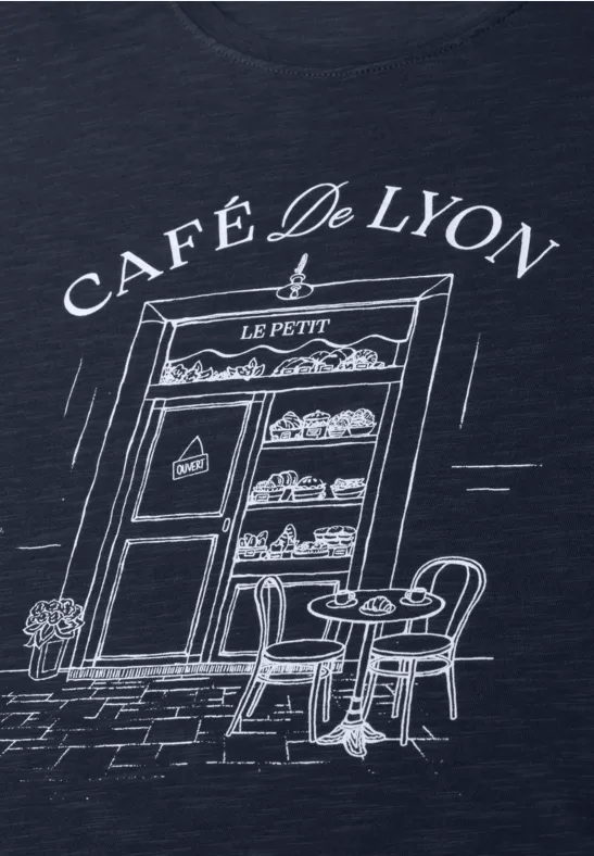 T-Shirt mit skizzenhafter Café-Fassade 'Café de Lyon', Vitrine mit Gebäck und Straßentisch mit Stühlen.