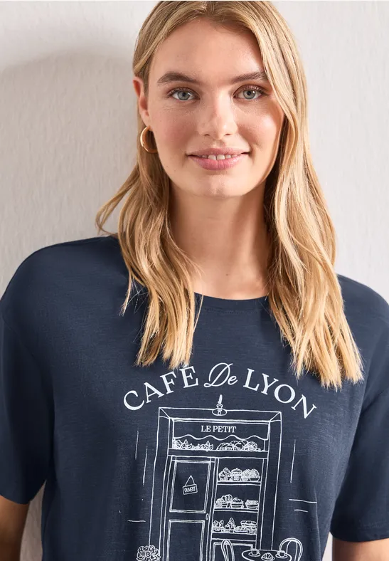 Frau mit blonden Haaren trägt ein navyblaues T-Shirt mit 'Café de Lyon' Aufdruck und einer skizzierten Bäckerei.