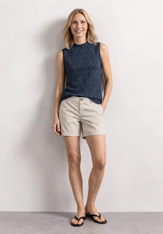 Frau in blauem Top und beigen Shorts mit Flip-Flops, vor hellem Hintergrund, lächelt entspannt.