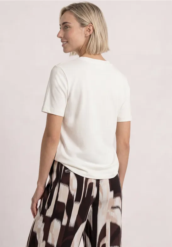 Frau mit kurzem blondem Haar trägt weißes T-Shirt und gemusterte, weite Hose, Rückenansicht.
