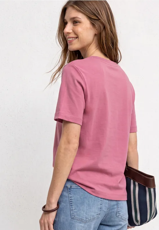Frau mit rosa T-Shirt und Jeans, lächelnd mit gestreifter Tasche in der Hand, steht vor hellem Hintergrund.