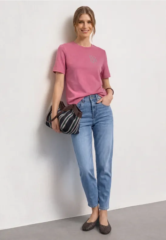 Frau in pinkem T-Shirt, blauen Jeans und Loafers, hält gestreifte Tasche vor weißem Hintergrund.