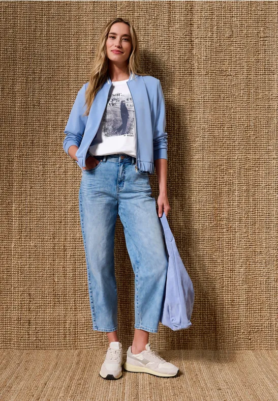 Vrouw in streetstyle: T-shirt met print, licht bomberjack, wijde cropped jeans en witte sneakers.