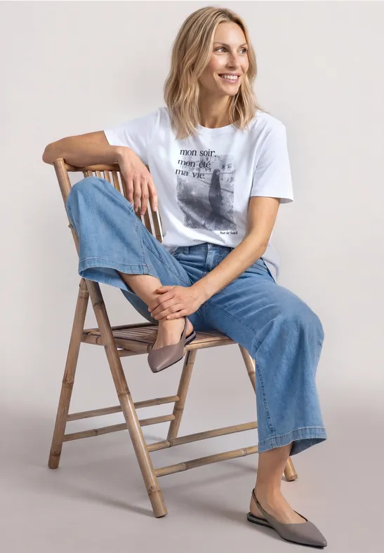 Een vrouw zit ontspannen op een houten ladder, draagt een wit t-shirt, wijde spijkerbroek en taupekleurige schoenen.