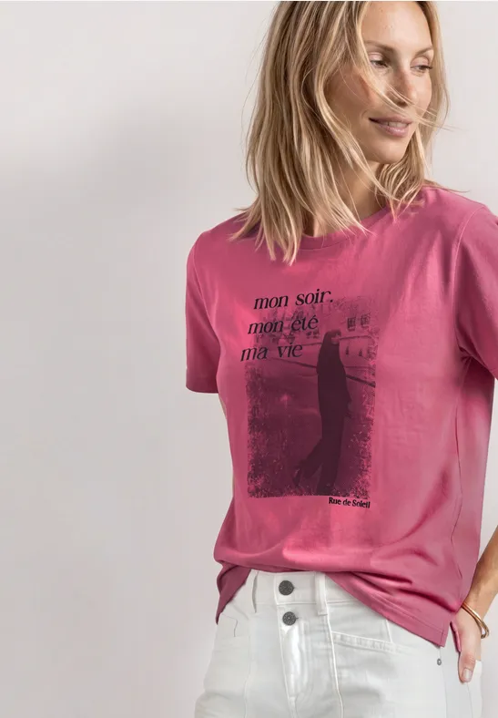 Eine lächelnde Frau in pinkem T-Shirt mit Aufdruck und weißer Hose, Blick nach rechts.